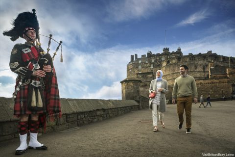 Couple walking towards man wearing Highland Dress, playing bagpipe with Edinburgh Castle, Scotland, in the background.